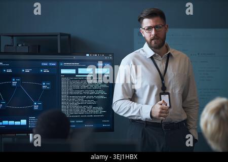 Medium shot of Caucasian young adult businessman presenting network analytics dashboard during office briefing, explaining cybersecurity data. Ideal for IT training, SaaS marketing Stock Photo