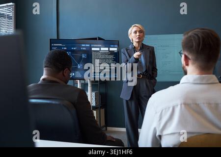 Caucasian middle aged businesswoman presenting software dashboard to colleagues during office meeting, explaining data workflow and strategy. Ideal for corporate training, leadership, teamwork Stock Photo