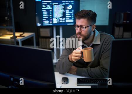 Caucasian young adult man monitoring code on computer screens while drinking coffee in office, representing software development and IT workflow. Medium shot, ideal for tech staffing or cybersecurity Stock Photo