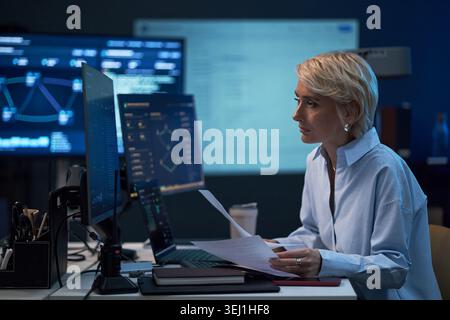 Middle aged Caucasian businesswoman reviewing documents and monitoring data dashboards on computer screens in office. Ideal for business analytics, finance strategy, cybersecurity Stock Photo