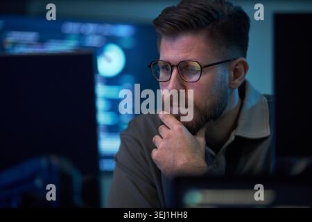 Close up of Caucasian young adult man analyzing data on computer monitors, touching chin while monitoring cybersecurity workflow. Ideal for IT security, analytics, remote work Stock Photo