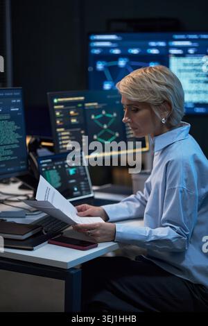 Vertical shot of middle aged Caucasian woman reviewing printed report beside cybersecurity dashboards and code screens, supporting IT audit, data security, compliance marketing Stock Photo