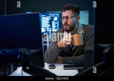 Caucasian young adult man monitoring code on desktop screens while drinking coffee during late shift, representing cybersecurity work. Ideal for IT staffing, software services, tech support Stock Photo