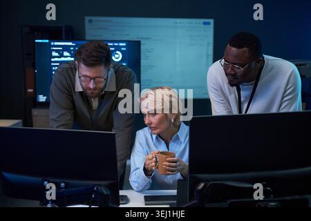 Medium shot of diverse business team analyzing data on computer monitors in control room, collaborating on problem solving. Ideal for cybersecurity, IT operations, teamwork marketing Stock Photo