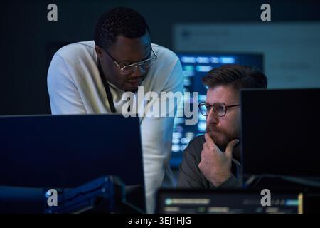 African American man and Caucasian man reviewing code on desktop monitors, collaborating on problem solving in IT office. Ideal for cybersecurity, software development, teamwork marketing Stock Photo