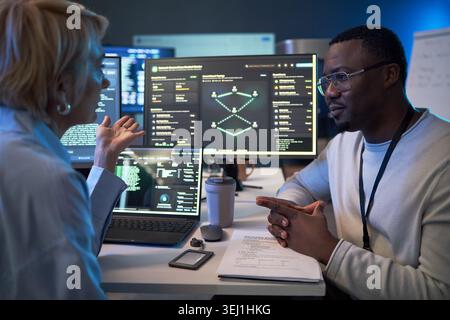 African American young adult man discussing cybersecurity data with middle aged Caucasian woman at workstation. Supporting IT consulting, software development, team strategy Stock Photo