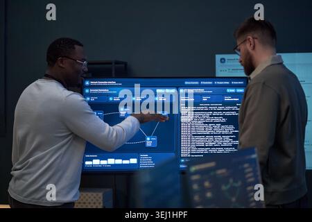 African American young adult man and Caucasian young adult man reviewing network dashboard, discussing code and system data. Useful for cybersecurity, IT teamwork, software development Stock Photo