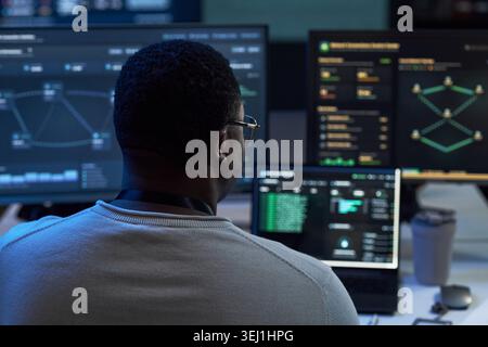 Rear view of Black young adult man monitoring cybersecurity dashboards across multiple screens, analyzing network threats and data flows. For IT security, SOC, risk management marketing Stock Photo