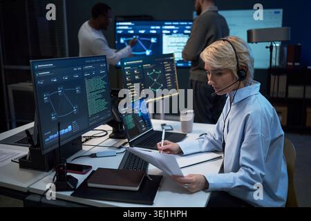 Caucasian middle aged woman wearing headset, reviewing reports and monitoring cybersecurity dashboard at workstation while colleagues analyzing data. For IT operations, network security, SOC marketing Stock Photo