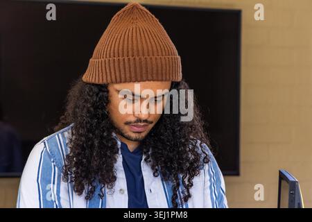 Adult male reading from monitor at home workspace, wearing beanie and striped shirt. Man, focus, screen, hat, knit, navy, brick Stock Photo