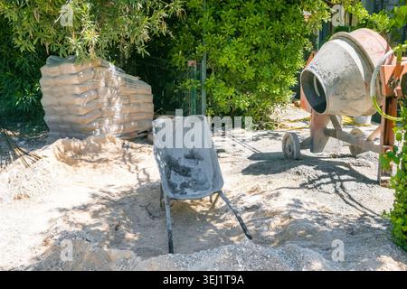 Mini concrete mixer and construction wheelbarrow on a construction site. Surrounding materials include sand and stacked bricks. Green foliage in the b Stock Photo