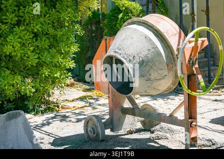 A mini concrete mixer sits on a construction site. It is orange and gray, with a partially filled drum. Surrounding debris indicates ongoing repair wo Stock Photo