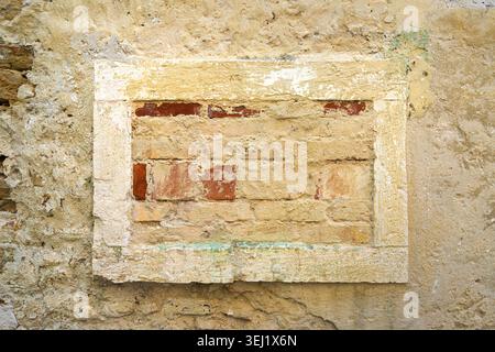 A section of weathered brickwork is set within a rough stone frame against a crumbling plastered wall. The bricks are unevenly laid and appear aged. Stock Photo