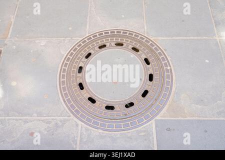 Piran, Slovenia. May 26, 2026.  A circular metal manhole cover with an ornamental design is centered on a tiled sidewalk. The manhole cover has a conc Stock Photo