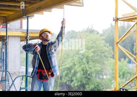 Construction worker wearing hardhat and safety glasses, managing a cable on a building site in rainy weather Stock Photo