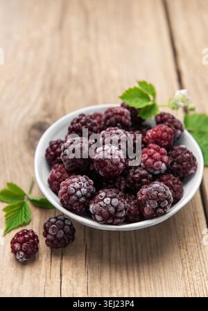 Frozen blackberries covered in frost filling a white bowl on a rustic wooden table Stock Photo