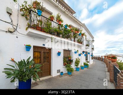 A charming street view featuring a whitewashed house facade with a plant-filled balcony and blue wall pots under a cloudy sky, Mijas, Andalusia, Spain Stock Photo