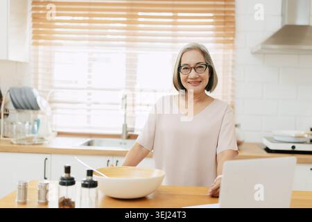 Senior Asian woman using Laptop computer modern technology to learn and live teaching cooking in the kitchen at home happy smile Stock Photo