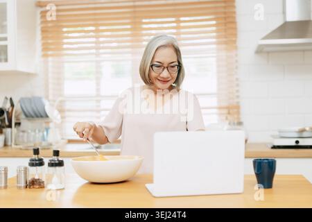 Senior Asian woman using Laptop computer modern technology to learn and live teaching cooking in the kitchen at home happy smile Stock Photo