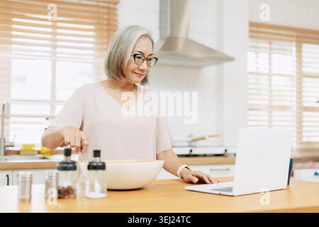 Senior Asian woman using Laptop computer modern technology to learn and live teaching cooking in the kitchen at home happy smile Stock Photo