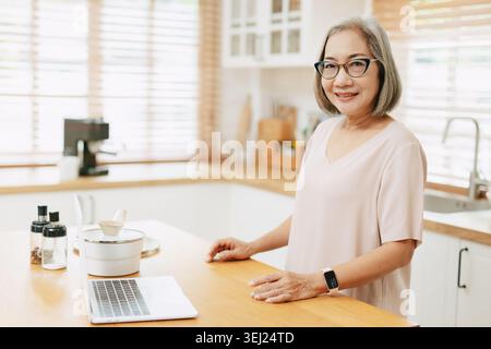 Senior Asian woman using Laptop computer modern technology to learn and live teaching cooking in the kitchen at home happy smile Stock Photo