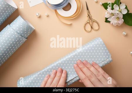 Top view of woman's hands wrapping a gift in blue polka dot paper, surrounded by gold scissors, ribbons, and flowers on a beige background. Stock Photo