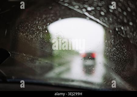 A point of view shot from inside a vehicle looking through a water covered windshield with selective focus on raindrops, showing a blurry car ahead an Stock Photo