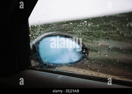 A close-up view from inside a vehicle looking through a wet side window covered in raindrops, showing an out-of-focus car wing mirror during bad weath Stock Photo