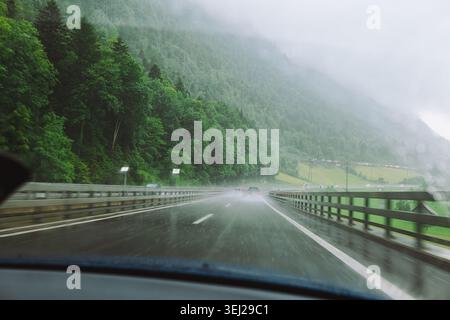 A point of view shot from inside a vehicle driving along an asphalt highway during a rainstorm, showing a wet road surface, traffic ahead, and lush fo Stock Photo