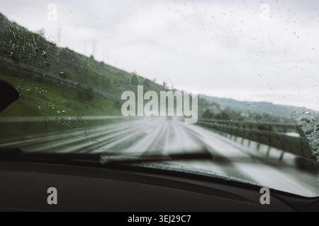 A view from inside a car dashboard looking through a rain covered windshield with water droplets on a cloudy day, showing an asphalt road and green hi Stock Photo
