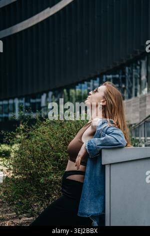 Woman wearing sunglasses tilts head up while leaning against outdoor structure in corporate district. Stress management metropolitan, taking a mindful Stock Photo