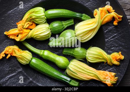 Fresh zucchini and blossoms arranged on dark plate, organic vegetables with edible flowers, cooking ingredient concept. Green squash and yellow blossoms top view Stock Photo