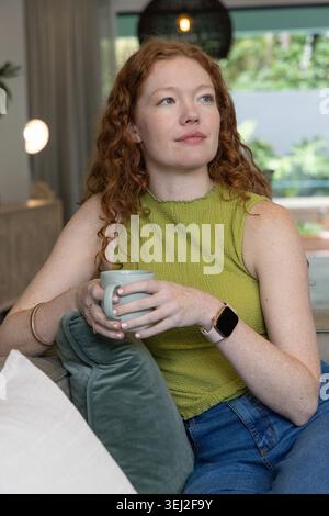 Red-haired woman sitting on sofa holding ceramic mug wearing green sleeveless top and jeans Stock Photo