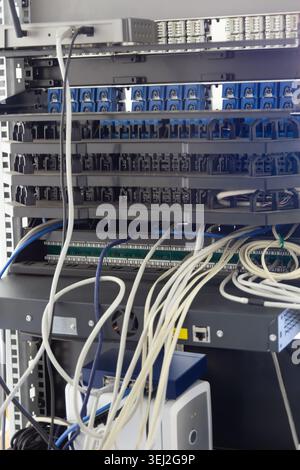 server rack containing network switches and devices with disorganized, tangled white and blue ethernet cables hanging in front Stock Photo