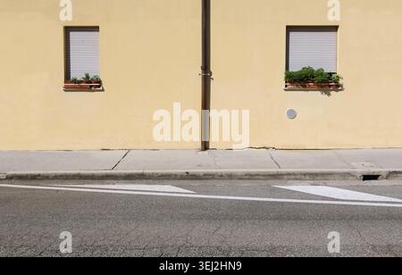 Brown roof gutter on yellow plaster wall with two closed windows by sides. Concrete sidewalk and street in front. Background for copy space. Stock Photo