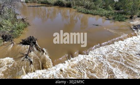 Aerial view of a muddy river flowing over a small weir with white foam and green vegetation on the banks Limpopo, Limpopo, South Africa. Stock Photo