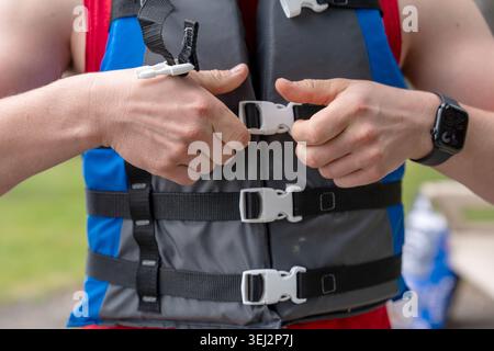 A close-up photograph of a fit man adjusting and buckling a blue and grey life vest (personal flotation device) outdoors. He is wearing a red tank top Stock Photo