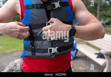 A close-up photograph of a fit man adjusting and buckling a blue and grey life vest (personal flotation device) outdoors. He is wearing a red tank top Stock Photo