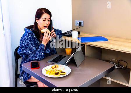 Woman multitasking working remotely eating breakfast talking on phone Stock Photo