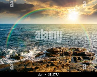 sunset over sea coast with stones and boulders. view of beach with rock in seaweed. dynamic cloudscape on the sky with rainbow above horizon afternoon Stock Photo
