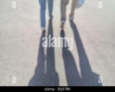 Two People Casting Long Blurred Shadows While Walking On Sunlit Pavement Producing Elongated Silhouettes And Abstract Ground Patterns In Urban Outdoor Setting With Overhead Light. Stock Photo