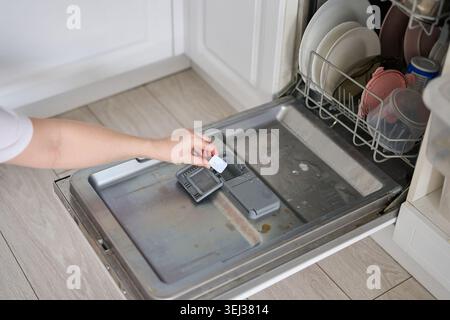 Side View Of Person Placing Dishwasher Detergent Tablet Into Dispenser On Open Dishwasher Door In White Kitchen Interior With Visible Cabinetry Plates And Tiled Flooring. Stock Photo
