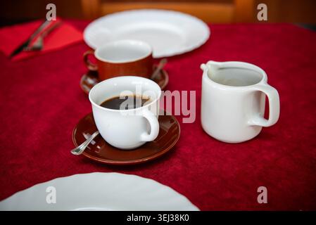 Cozy dining table prepared for breakfast or brunch with coffee cups, white plates and red tablecloth in a warm rustic interior. Inviting restaurant or Stock Photo