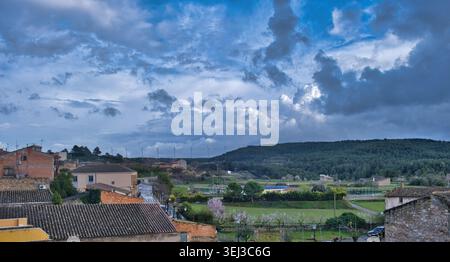 Dark clouds gather above a Spanish village with tiled roofs, green fields, and wind turbines on a distant hill. Panoramic view at Fulleda,  Catalonia Stock Photo
