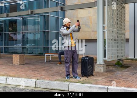 Holding man checking smartphone at plaza, wearing beanie and denim jacket, with wheeled suitcase Stock Photo