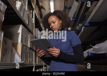 A female warehouse worker checks inventory while organizing packages in an industrial space focused on logistics using tablet. Stock Photo