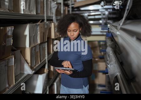 Woman managing stock and organizing inventory on shelves in a busy warehouse using digital tablet. Stock Photo