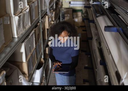 Female warehouse worker checks inventory on a tablet while organizing packages in an industrial storage area. Stock Photo