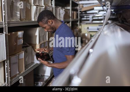An African American man checks inventory and organizes packages in a warehouse with digital tablet. Stock Photo