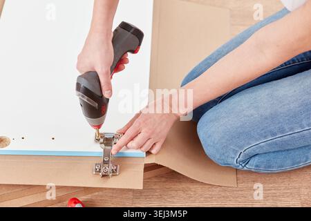 A person is sitting on the floor, using a cordless drill to attach hinges to a cabinet door in a well-lit, inviting space filled with wooden elements Stock Photo